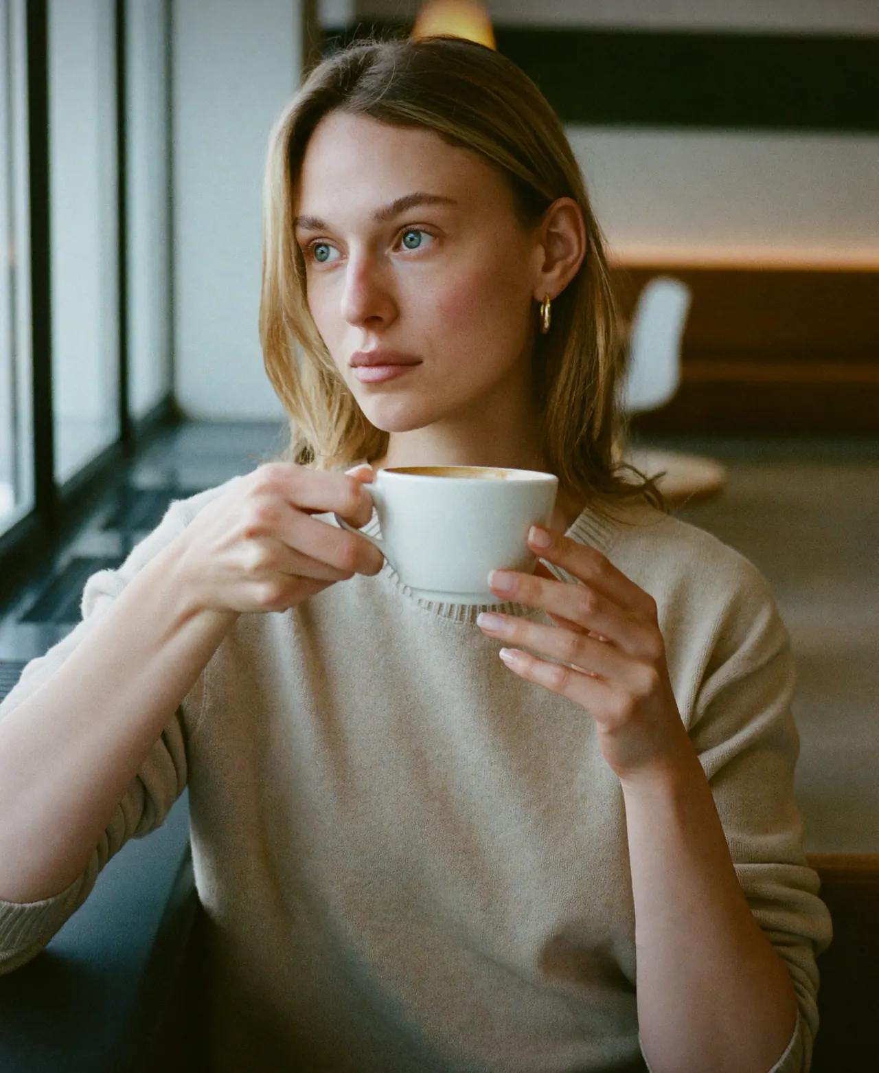Image au format créateur de contenu, une jeune femme tient une tasse à contre-jour devant la baie vitrée d'un café, lumière du lieu uniquement